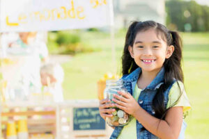 A small girl with a jar of coins in front of a lemonade stall showing the idea of entrepreneurship in school kids
